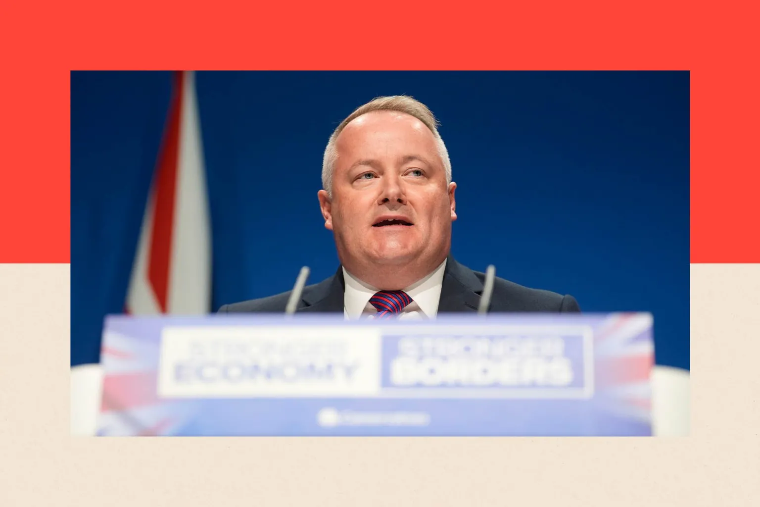  Darren Millar, the Conservatives' Senedd leader, stands in front of a blue backdrop and a union flag. In front of him is a lectern with a microphone. The words 