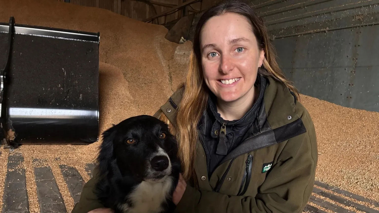 Anna Jackson is wearing a green jacket and kneeling next to her black and white boarder collie dog. They are in a grain store and a large pile of grain can be seen behind them.