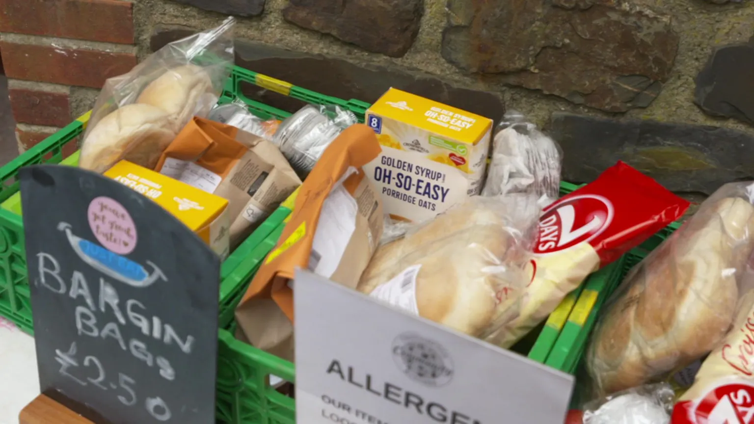 Two green boxes containing food, including instant porridge, bread and all sorts of different packets of pastries. There is a blackboard sign and it reads: 'Bargain Bags £2.50'.