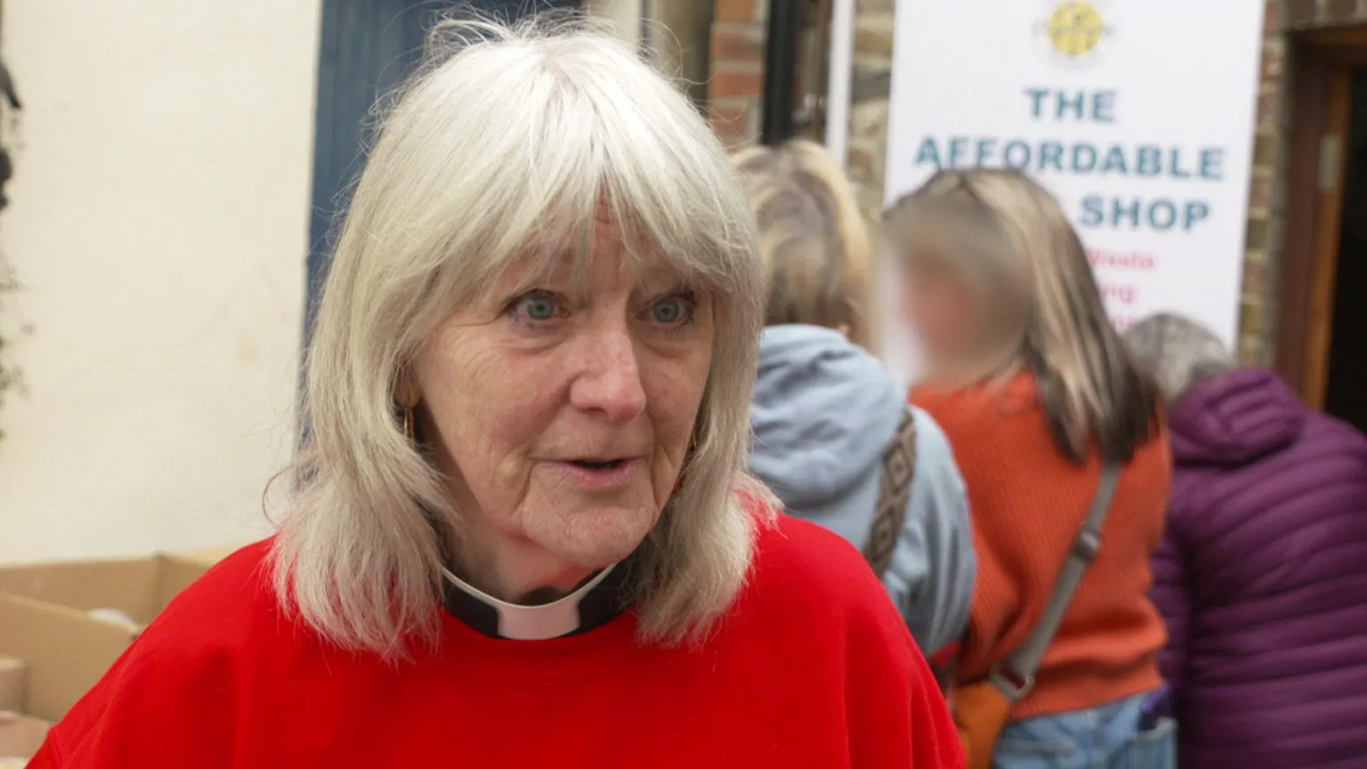 Older woman with shoulder length wavy hair, wearing a vicar's white collar and red sweatshirt.