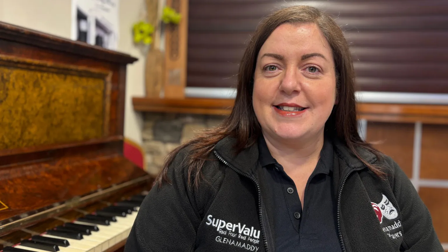 A woman sits beside a piano. She has long dark hair and is wearing a jacket with a theatre emblem on the chest. She is smiling. 