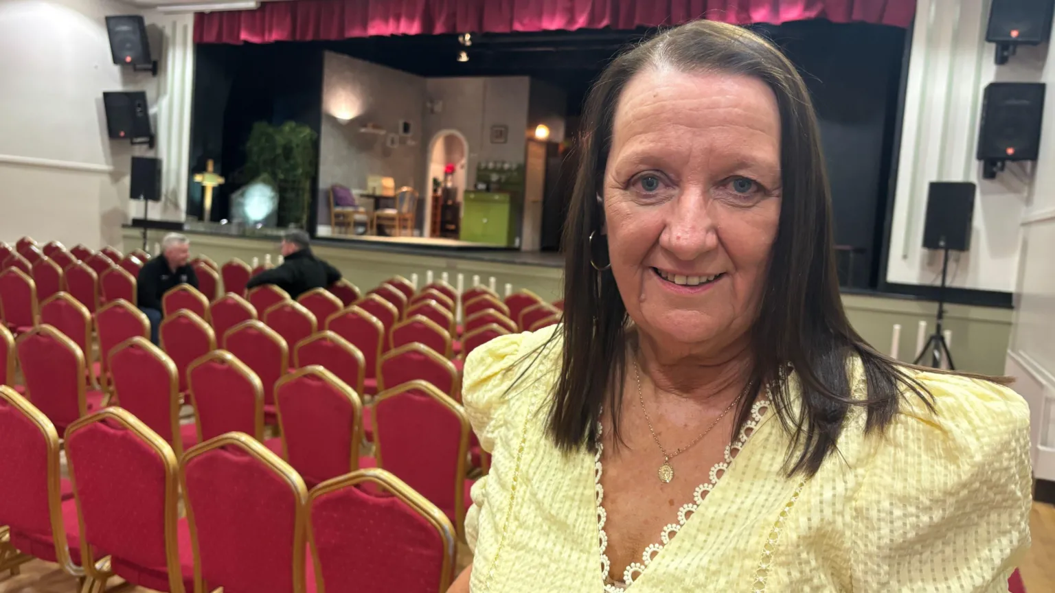 A woman in a yellow summer dress stands in a theatre. She has dark hair over her shoulders, and is smiling. Behind her are rows of red velvet seats with gold surrounds. The stage in the background is partially lit up as though a play is about to be staged. 
