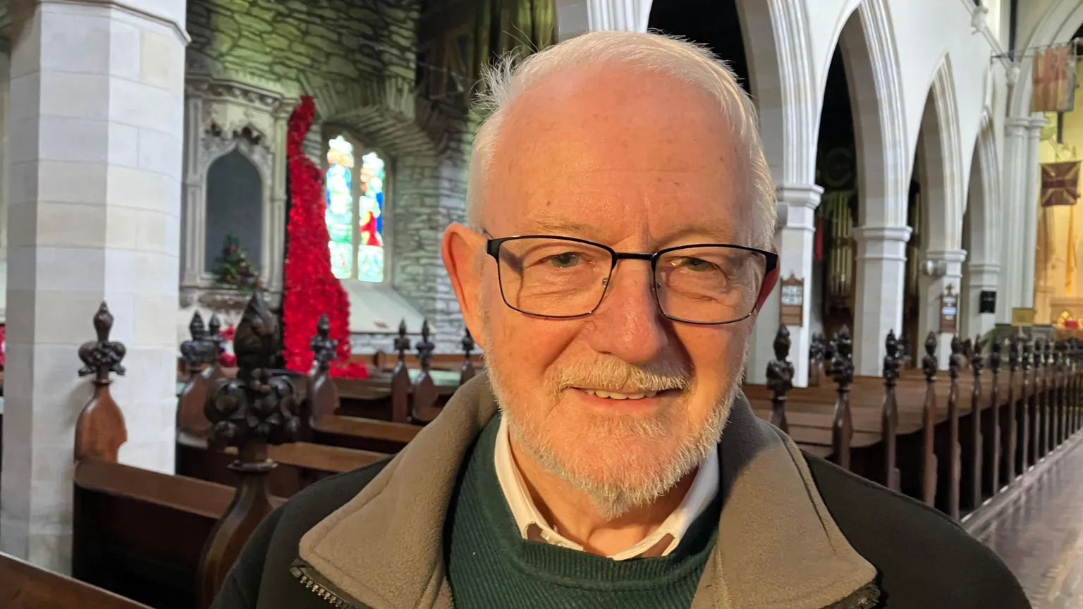 Robert wears spectacles with fine, rectangular frames. He is wearing a jacket unzipped, which shows the collar of his jumper and shirt. He is smiling. Behind him are the pews of St Columb's Cathedral. 
