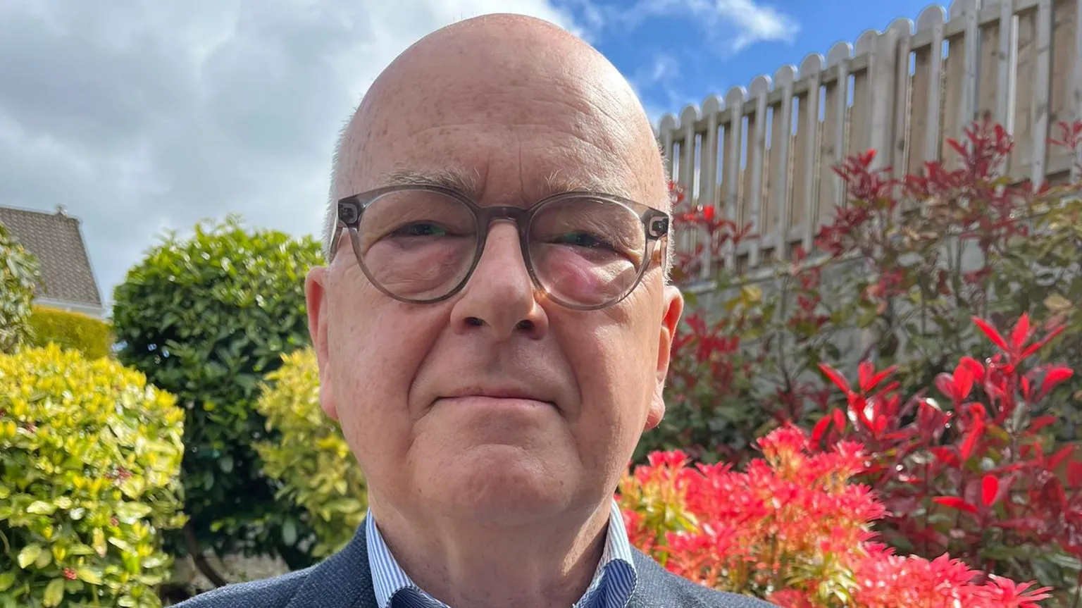 Drew is bald and bespectacled. His glasses a more round than they are square, and a shadow is cast over his restful face. Behind him are plants and shrubs. 