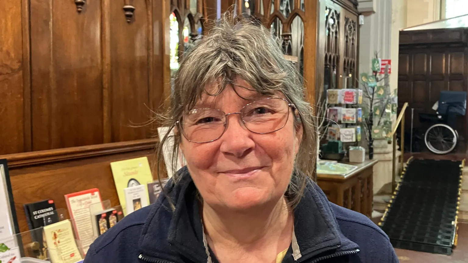 Jane has large glasses and messy grey hear. She is smiling and wearing a navy double-collared fleece. Behind her are religious pamphlets arranged along a shelf. 