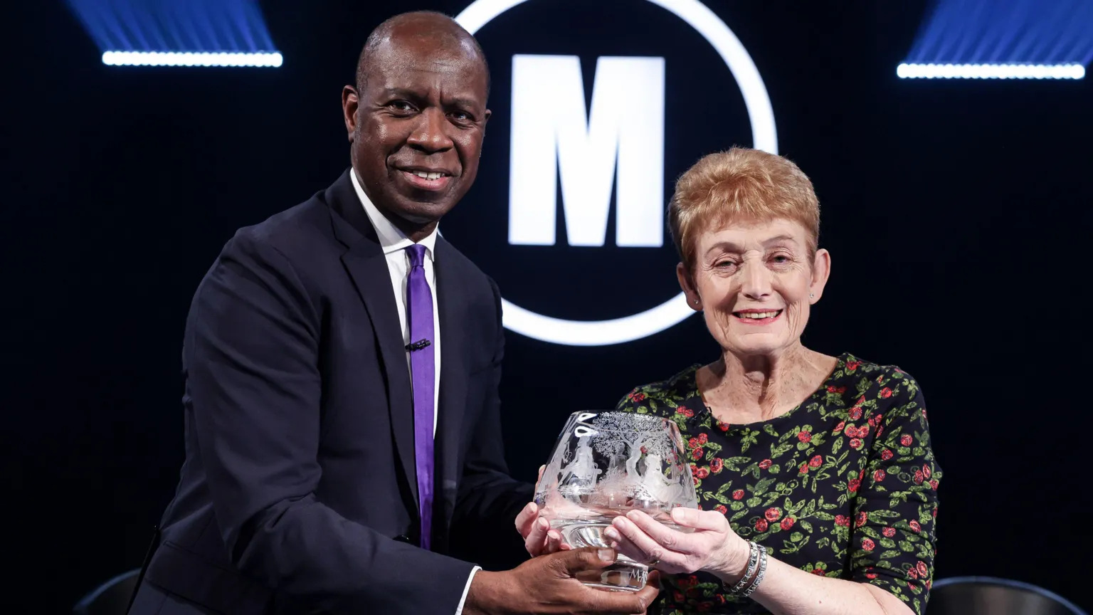HatTrick/BBC Diane with her trophy and show host Clive Myrie, who is wearing a suit with a purple tie.