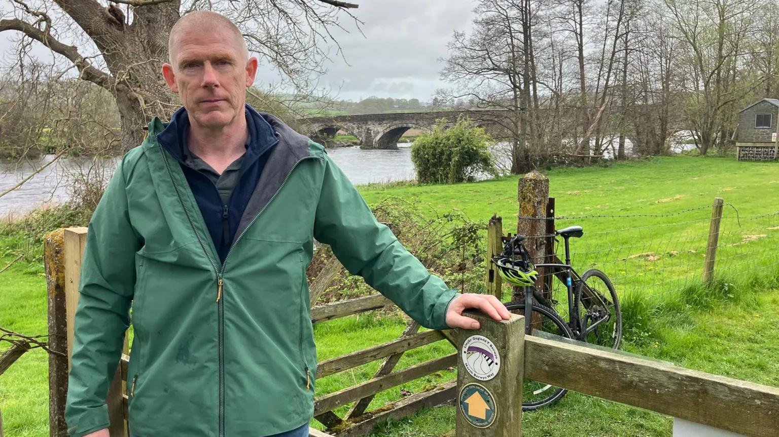 Dafydd Morris stands by the stile with his hand on the post with the trail marker. 