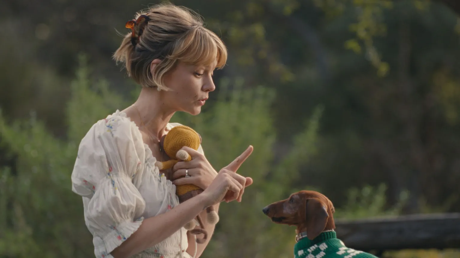 Netflix Carey Mulligan in Beef 2, wearing a white patterned dress and holding a finger up to to a dog, who is wearing a green jumper 
