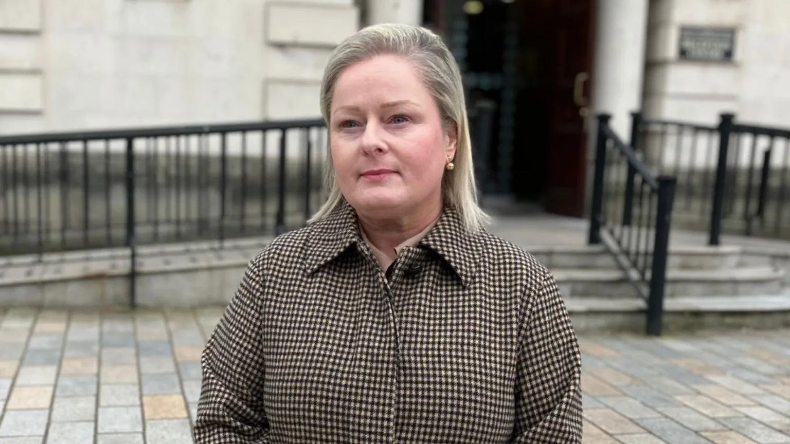 A woman with shoulder-length blonde hair wearing a brown and cream checked coat, stood outside the steps and railings of a courthouse
