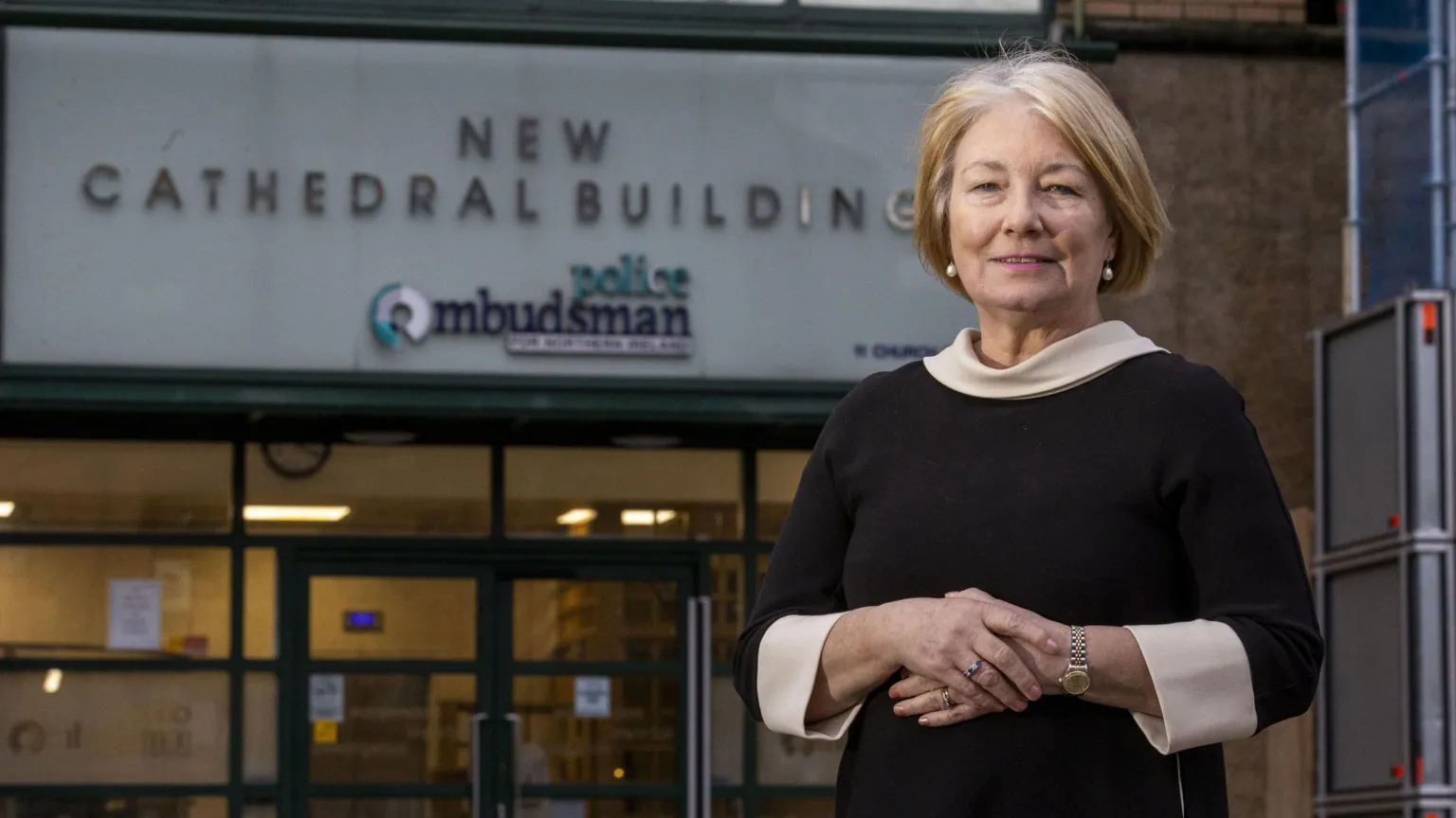 Liam McBurney/PA Wire Marie Anderson outside her office in Belfast. She has one hand resting over the other and has a ring on both hands. She's wearing a silver watch and peral earrings and a black and white dress. The sign in the background says new cathedral building, police ombudsman. 