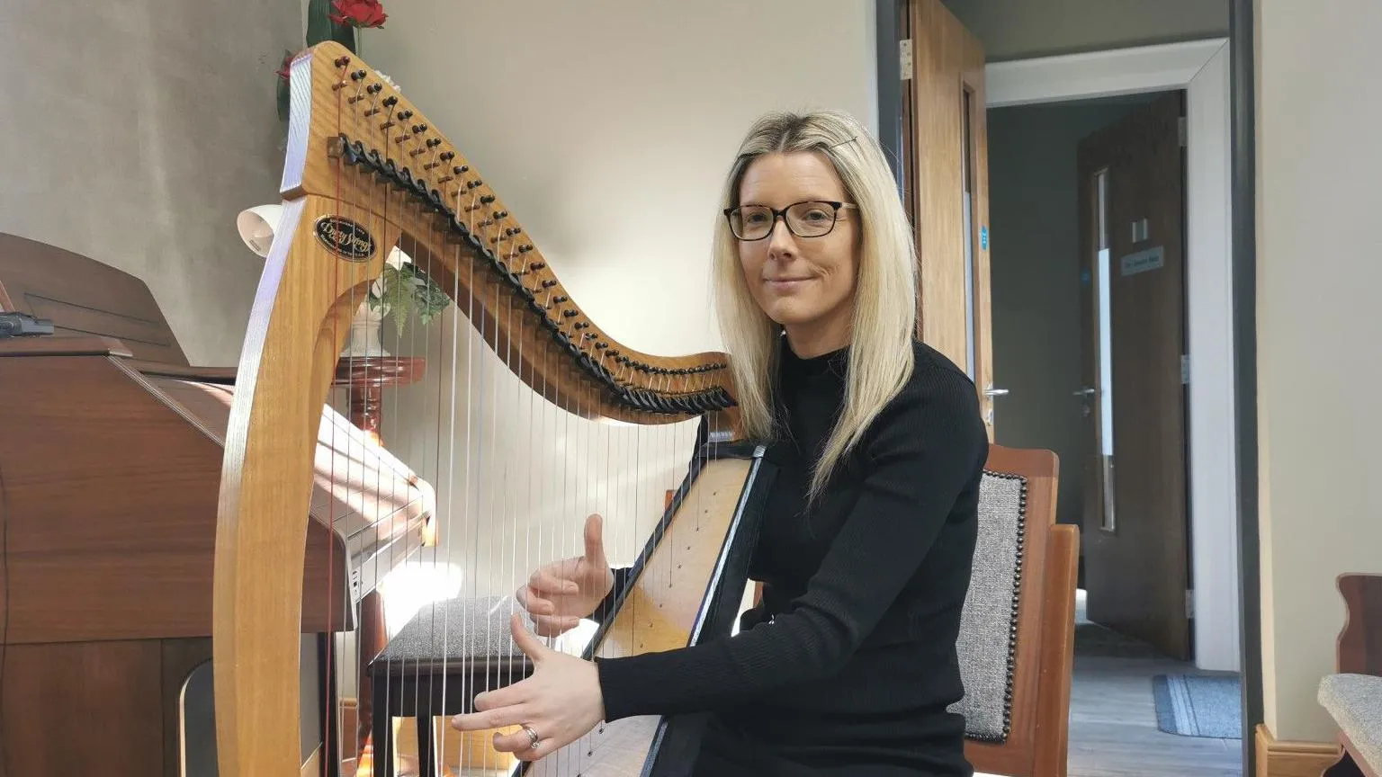 Clara Wilson is sitting in a church with a large harp. Her two hands are playing the strings. She's looking into the camera. She wears glasses and her hair is blond. She has black clothes on. An organ is beside her. 