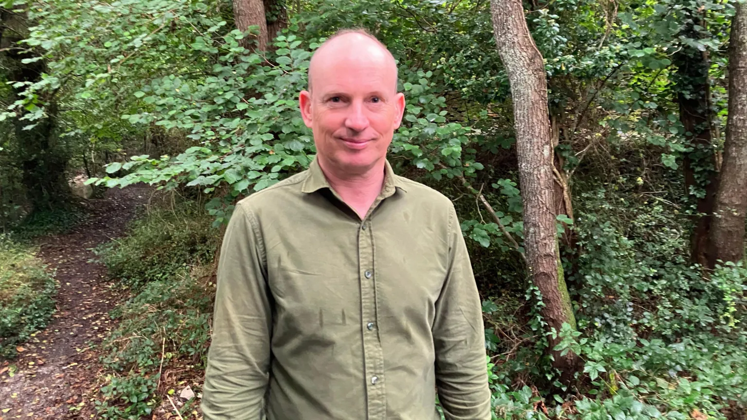 A man wearing a green shirt standing outdoors in a woodland area. 