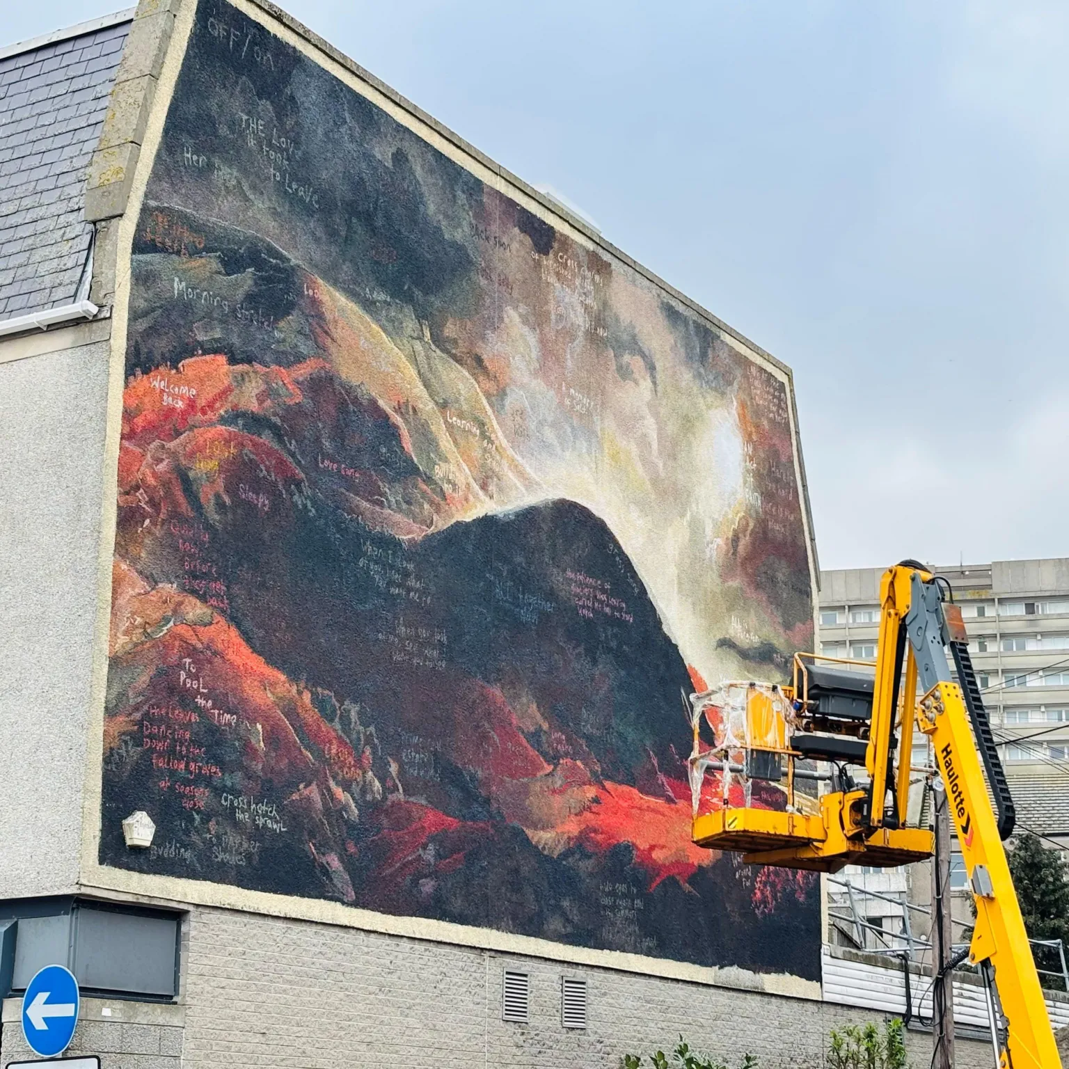 An arwork taking shape on a building wall, a yellow cherry-picker height vehicle is next to it, and a block of flats is behind.