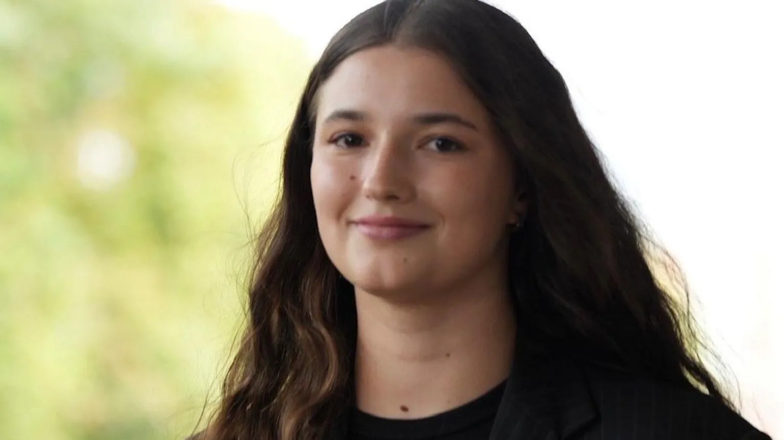 Oksana Lohvynenko A brunette girl stood in front of a nature backdrop, she smiles towards the camera as her hair flows in the wind. She is wearing a pin-striped black blazer with a rose attached to her side. 