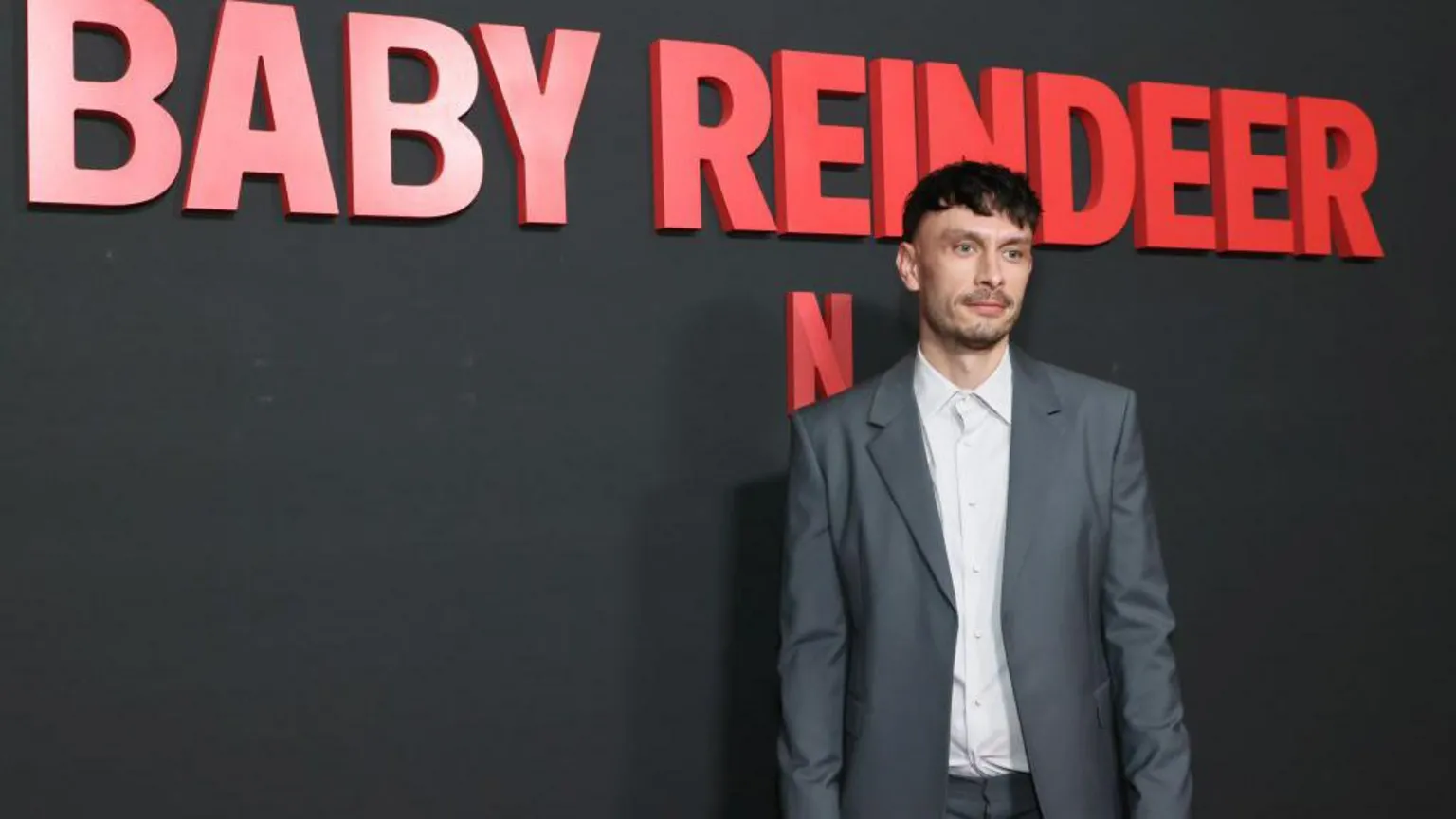  Richard Gadd, looking thin and pale in a white shirt and grey suit, in front of a grey wall and the name Baby Reindeer logo in red lettering.