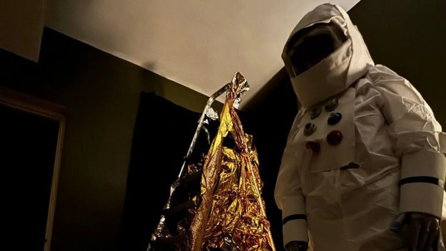 Deby McKnight A person wearing a white spacesuit and space helmet. There is a ladder behind them, which is wrapped in gold-coloured material. 