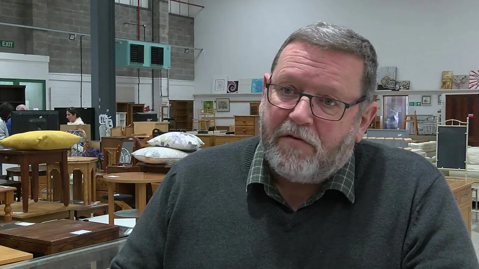 Kelvin Hughes is sat in a furniture showroom. He has short, greying hair and is wearing a green stripey shirt and a grey or black jumper.