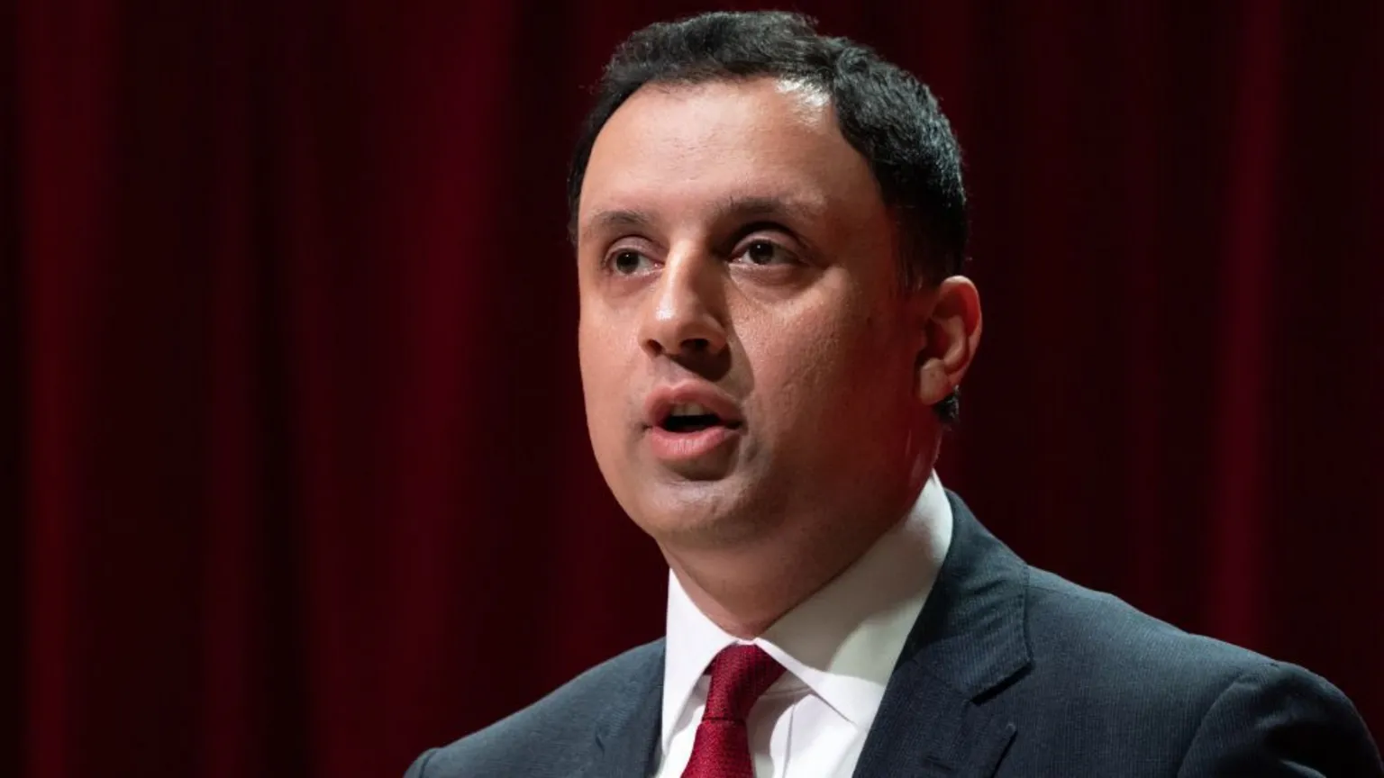  Anas Sarwar, who has short black hair, wearing a business suit and a red tie. There is a red curtain 
