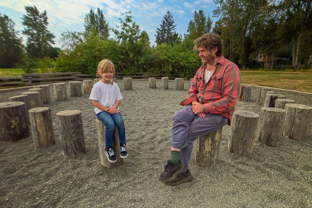 Zach Galifianakis wearing a red plaid shirt and grey trousers talks to a boy in white T-shirt and jeans as they sit on logs arranged in a circle