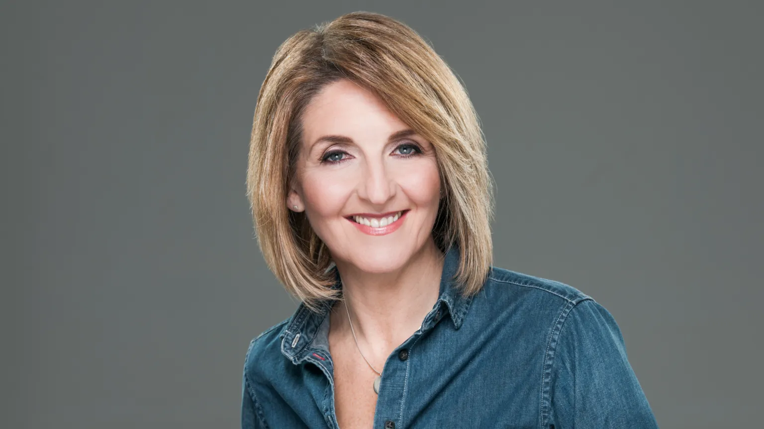 BBC Scotland Kaye Adams smiling straight at the camera. She is wearing a blue shirt and is standing against a grey background.