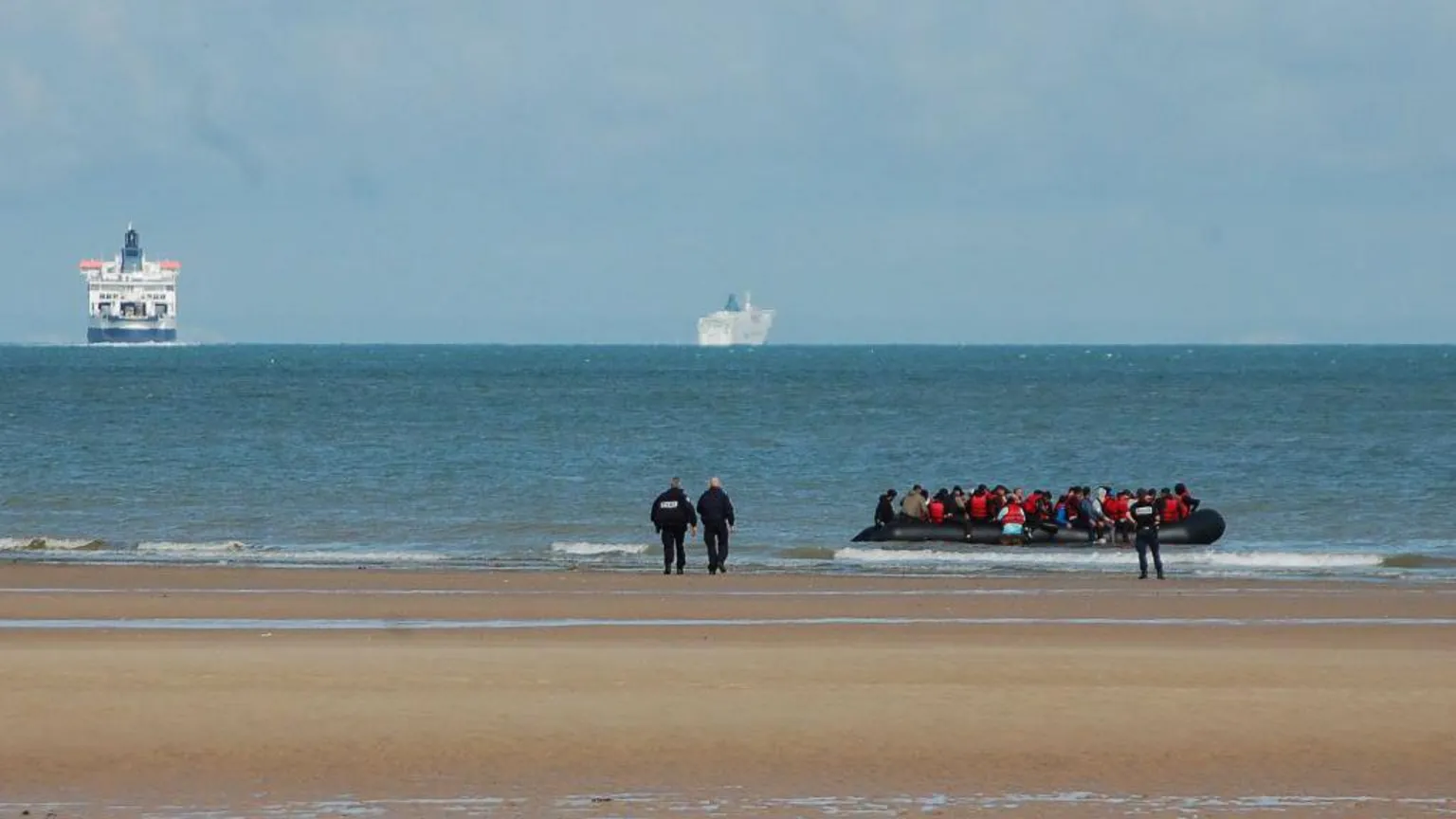BERNARD BARRON/AFP via Police officers on a sandy beach in France walk in the direction of an inflatable boat carrying migrants. In the distance are two ferries. 
