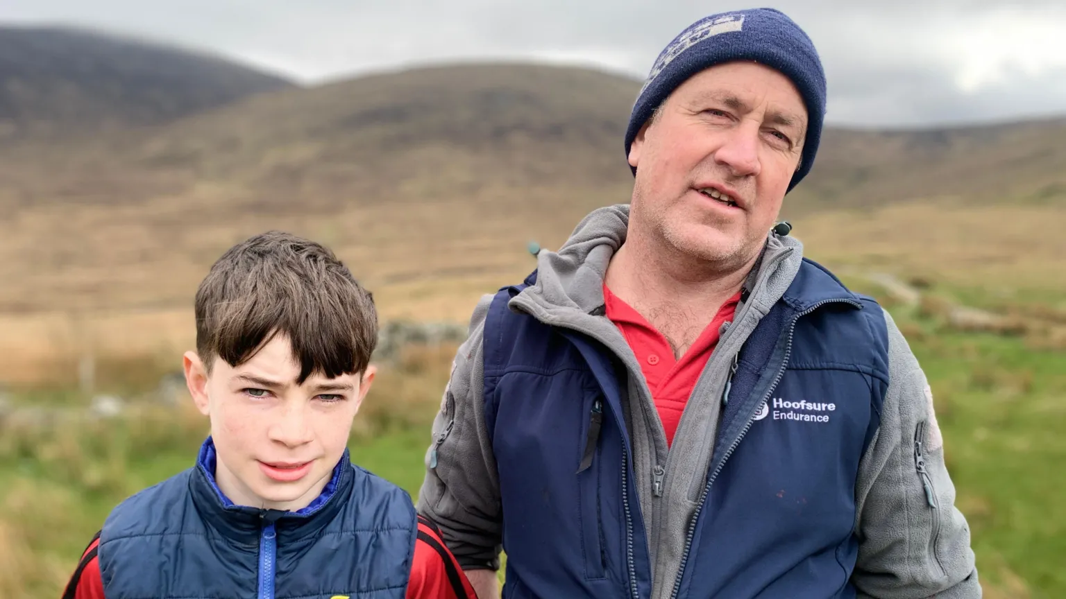 A young boy with brown hair and wearing a red t-shirt with a navy gilet over is is looking at the camera. He is on the left of the image. On the right is a man with a blue woolly hat on his head. He's wearing a red t-shirt, grey hoodie and navy gilet.