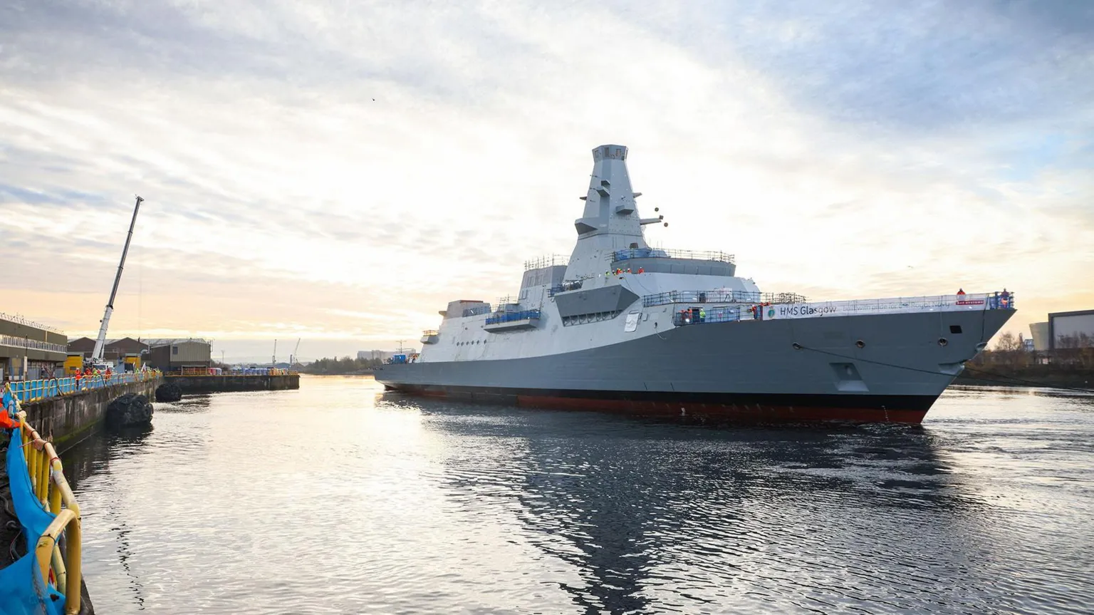 A large grey warship arriving at a shipyard on a river