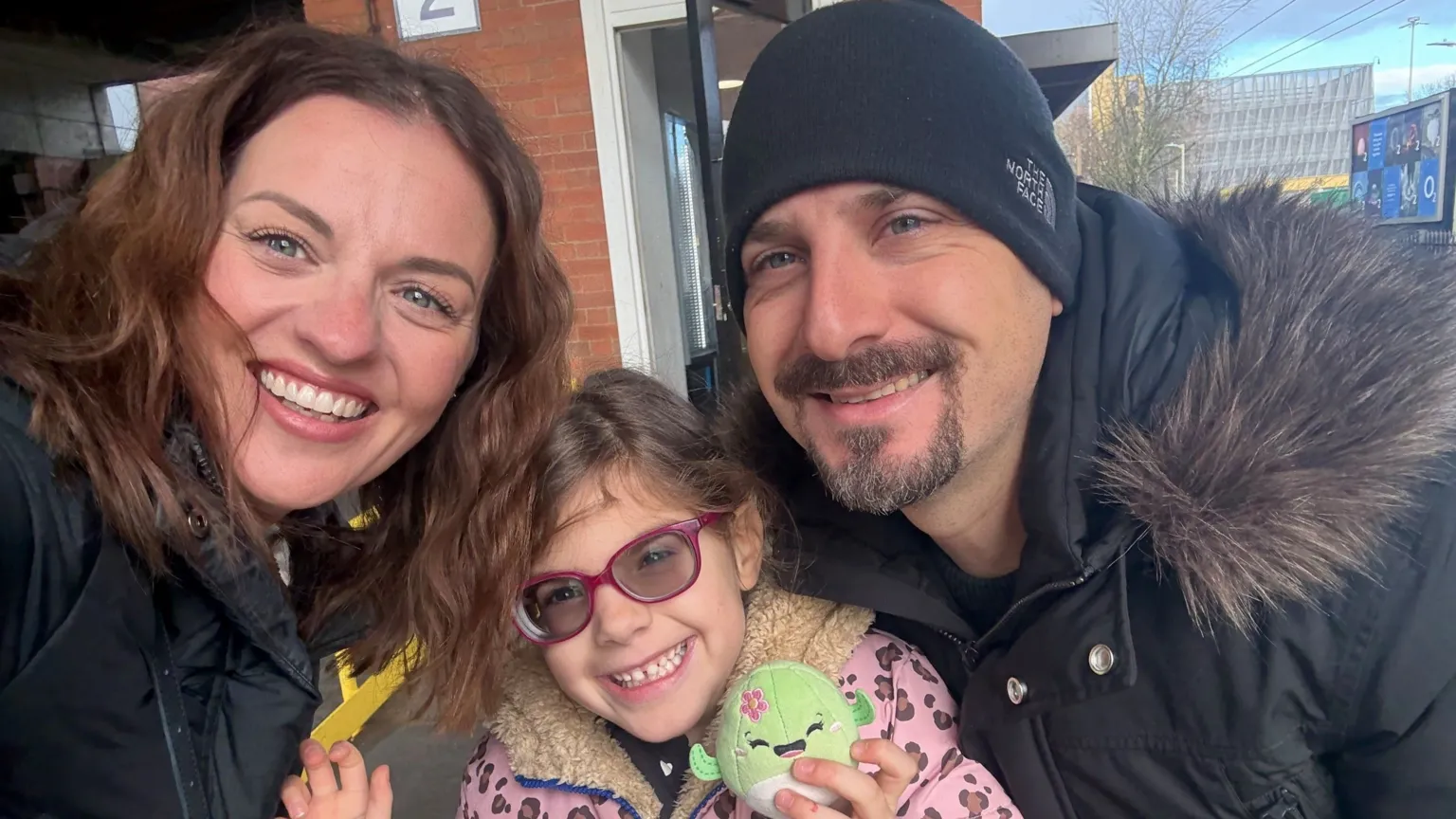 Family handout Three people all together smiling and looking at the camera. The young girl in the middle is holding a toy and has on a pink and brown leopard print coat, with purple glasses on. A woman is to the left, with long dark hair and a man to the right with a black hat on, and black coat with a furry hood. 