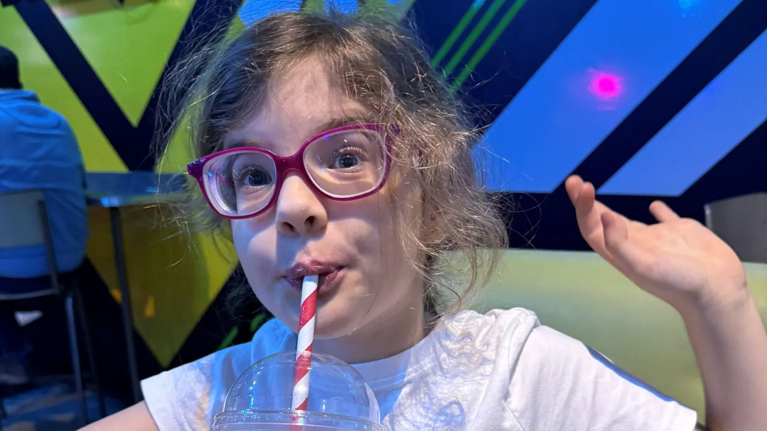 Family handout A girl, sucking a red and white stripped straw, in a drink. She has one hand up, and is wearing purple glasses and a white T-shirt. She is in a room that has bright colours on the wall behind her. 