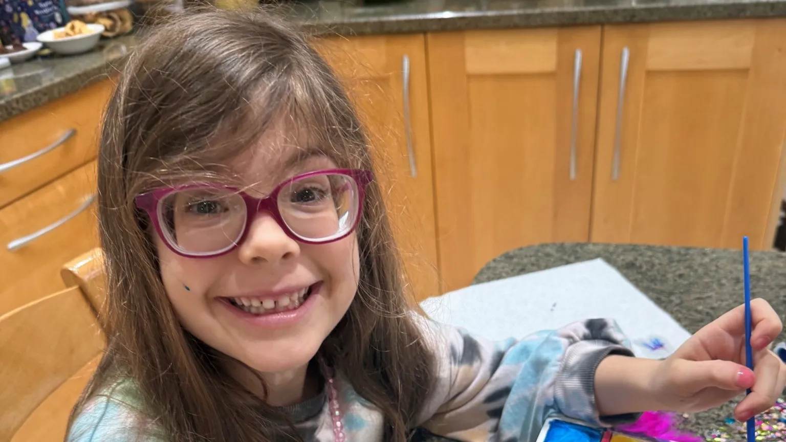 Family handout Saffie, wearing purple glasses, holding a paint brush in one hand. She has on purple glasses and has long dark hair. She is sat by a table in a kitchen, with cabinets all around her. 