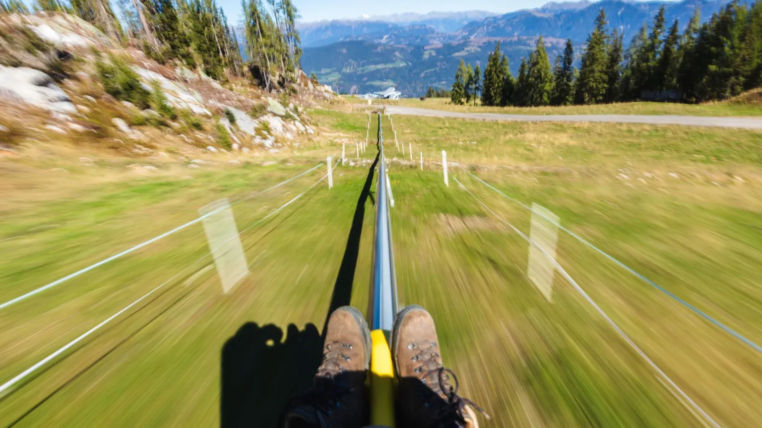  A person rides a yellow toboggan down a metal track on a mountainside in Germany. The person's brown walking boots is all that be seen of them. The grassy slope is slightly blurred as the toboggan slides down the rail. In the distance are pine trees and mountains. 