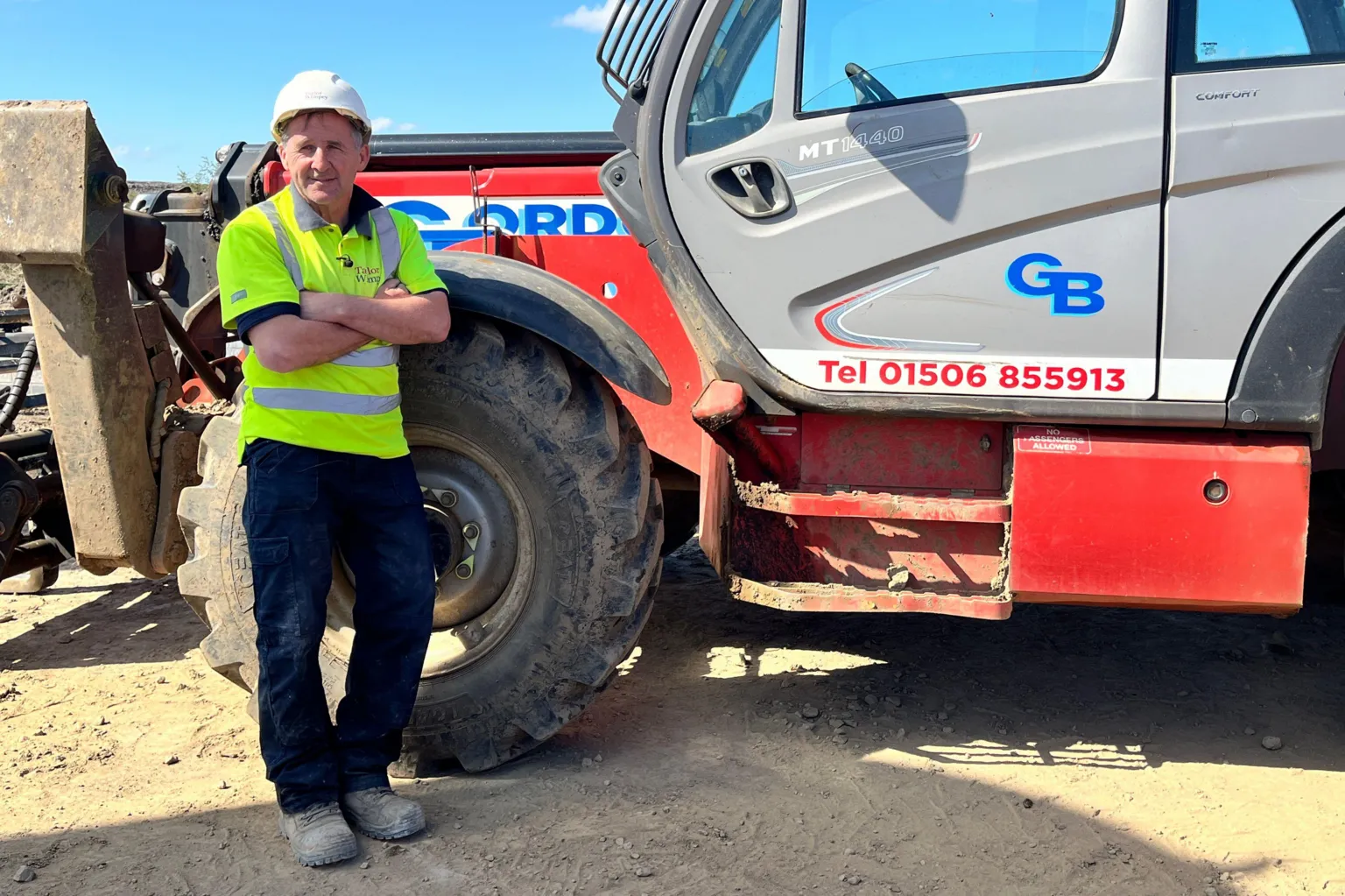 Construction worker in a high-visibility shirt and hard hat stands with arms crossed beside a large red and grey telehandler on a dusty worksite under a clear blue sky.