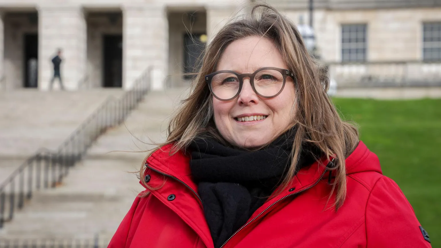 Cancer Research UK A woman with highlighted hair, glasses and red coat with black scarf stands in front of parliament building, stormont 