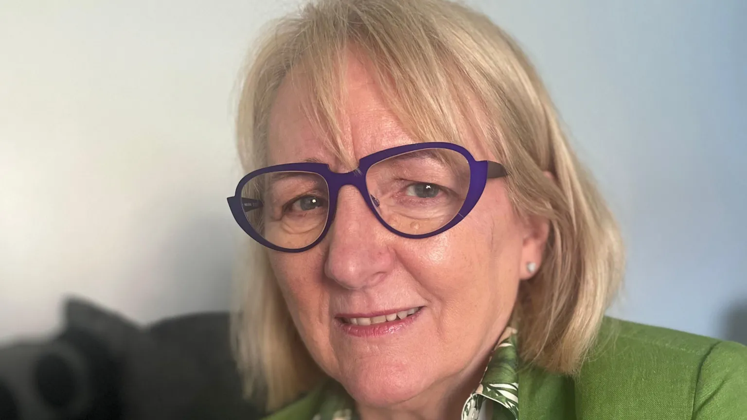 Jones has short blonde hair and a fringe. She is wearing purple glasses, a green blazer and patterned shirt.