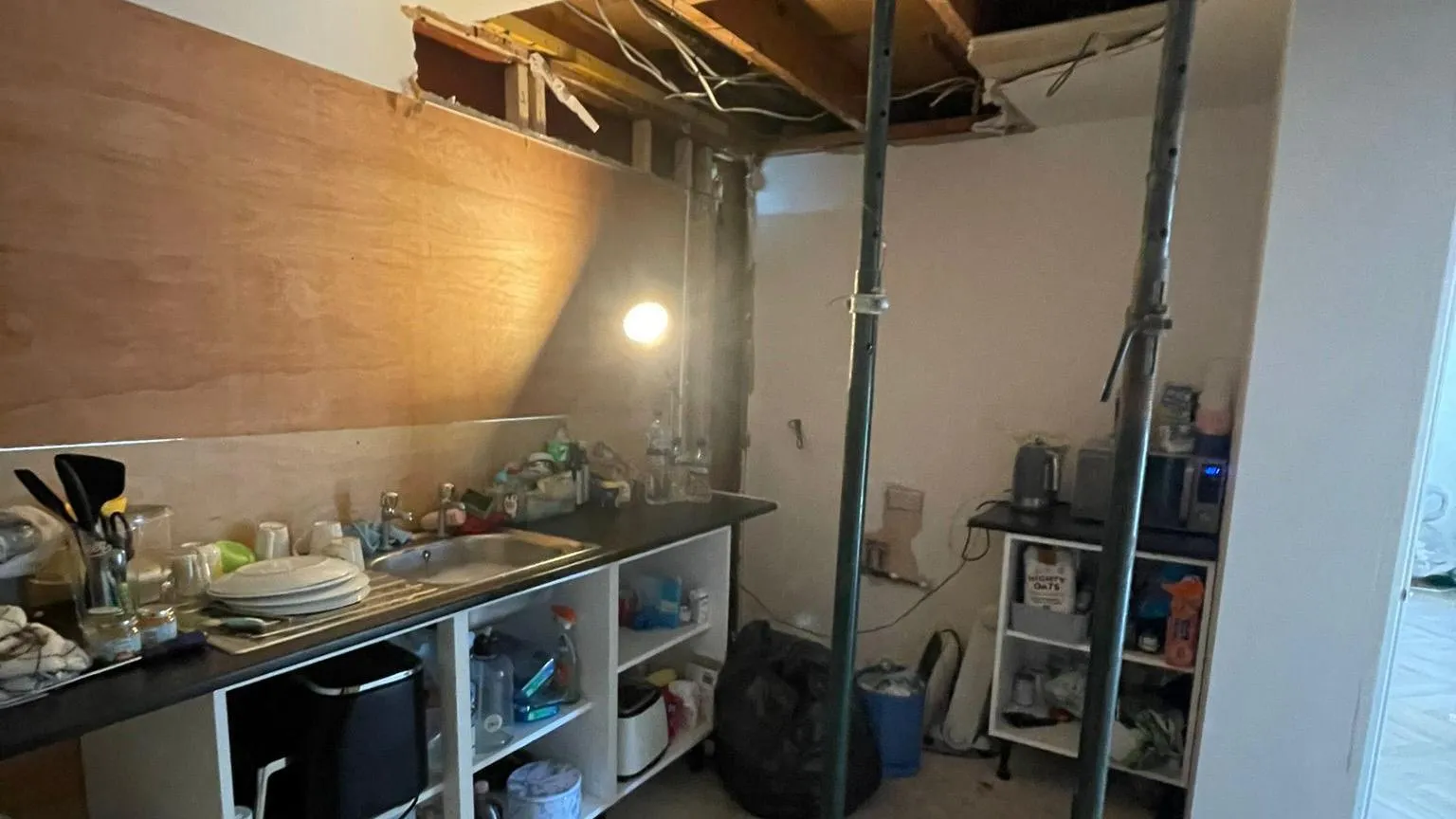 Rob Davies A kitchen with the ceiling being held up by tall black poles, but it is exposed and unfinished. There is a worktop with a sink and wires hanging out of a wall. It is a messy scene.