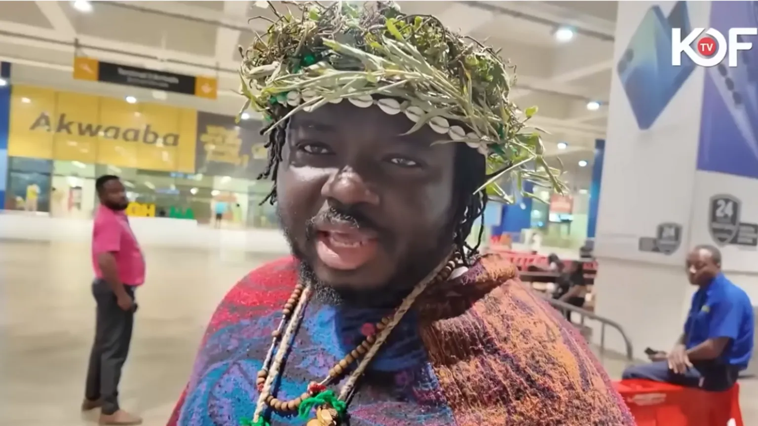 KOFI TV Screengrab of a video of tribesman in an airport. He is wearing a headdress made from green vegetation, and wooden necklaces. 