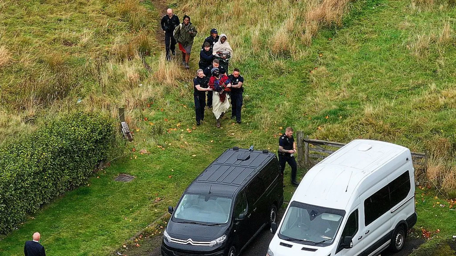  A drone shot of about seven uniformed officers escorting three people in robes along a grassy path. There are two vans waiting for them. 