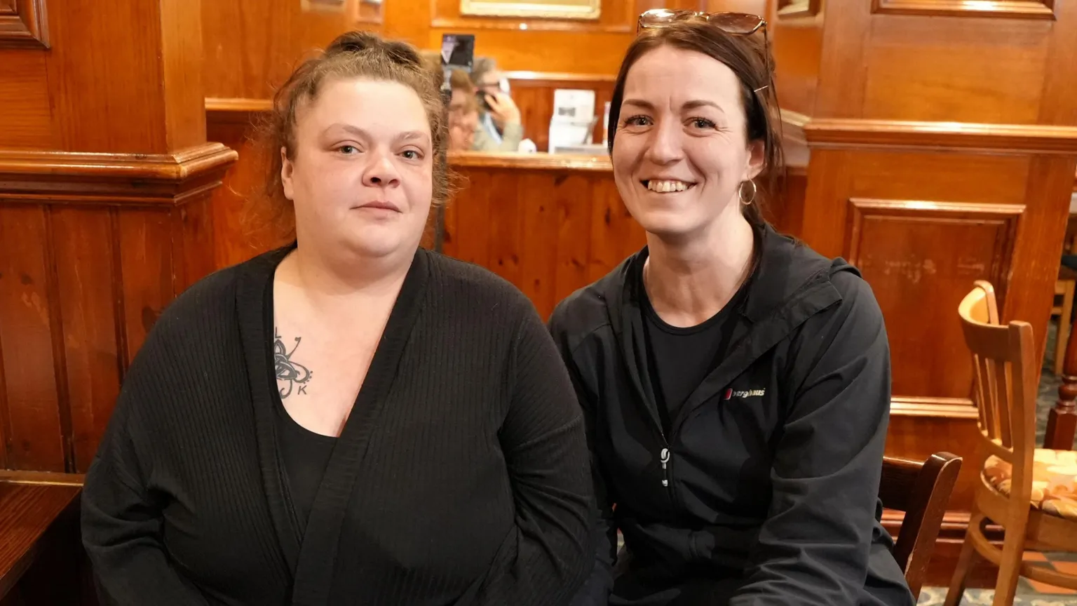 Kelsey Hesp (on the left of the photo) is wearing a black vest and black cardigan. Her brown hair is tied back. She is sat in a pub next to her friend, Natalie Wasiuk (on the right side of the photo) who is wearing a black top and black Berghaus jacket. Her hair is also tied back. She has sunglasses on her head.