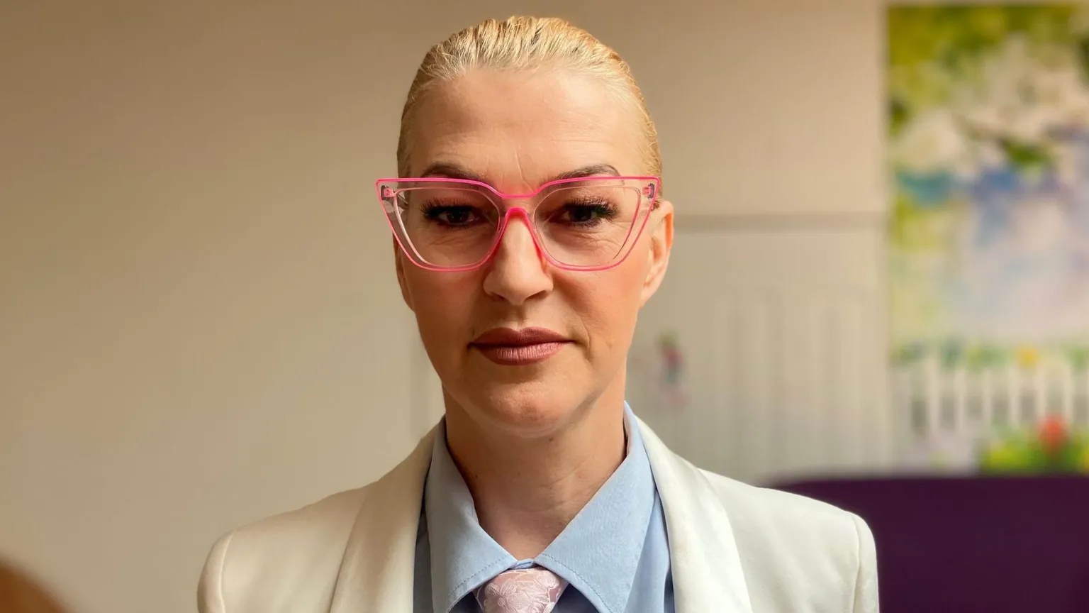 Anna Buckley's blonde hair is tied back. She is wearing pink glasses, a blue shirt with a pink tie, and a white blazer.