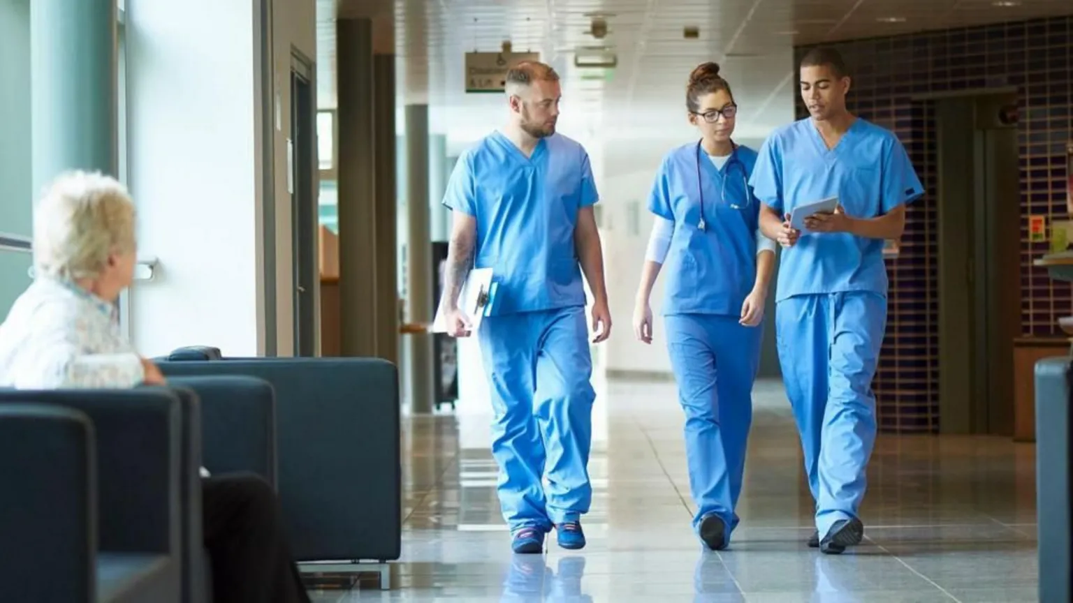  A stock image of three healthcare professionals walking through what appears to be a hospital corridor, dressed in blue scrubs. An elderly patient looks on from a seating area in the foreground.