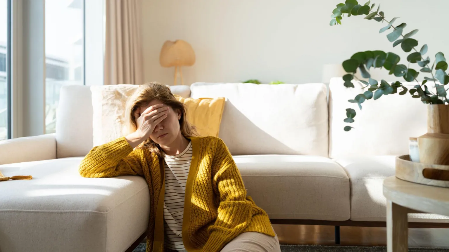  A stock image of a woman wearing a yellow cardigan and striped white t-shirt sitting on the floor next to a white corner sofa, with her hand covering her face in apparent discomfort. There is a green potted plant to the right of frame.