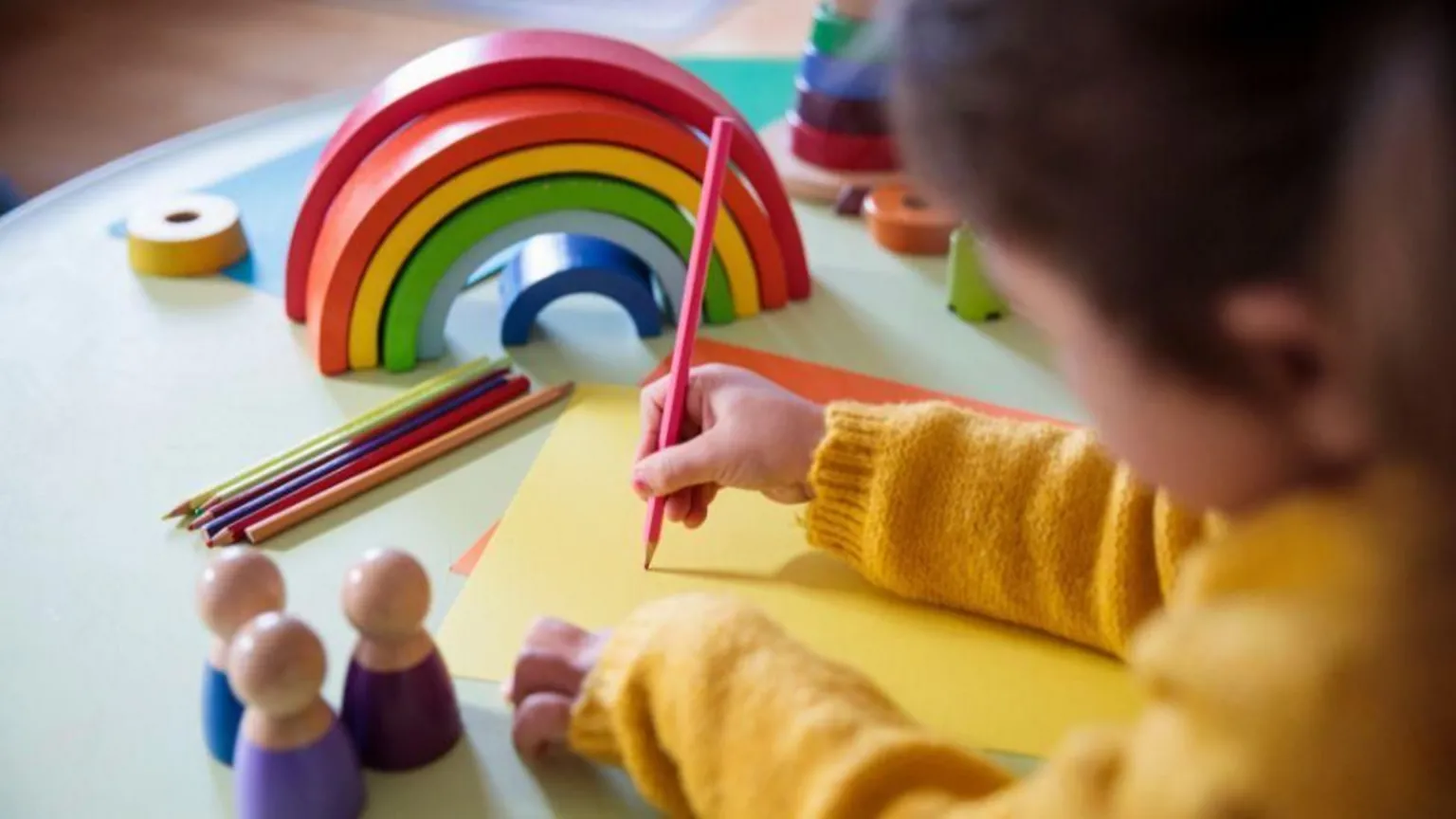  A stock image of a child wearing a bright yellow jumper, sitting at a table drawing with colourful pencils, with a wooden rainbow in the background. Their face is out of focus.
