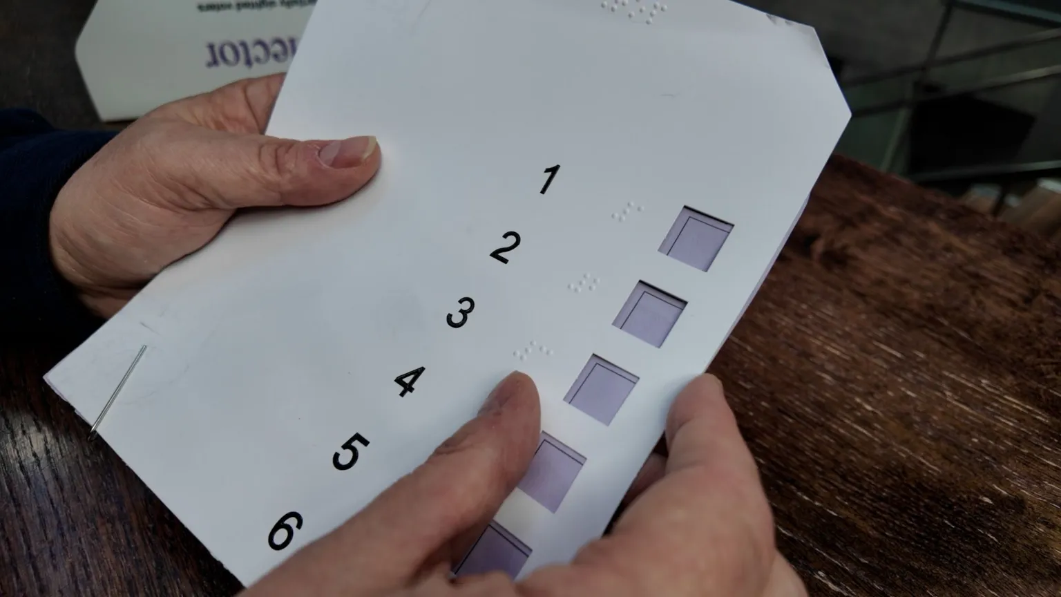A close up of two hands holding the voting template. It seems to be a thin plastic sheet with the numbers one to six printed from top to bottom and a small open square beside each one where 