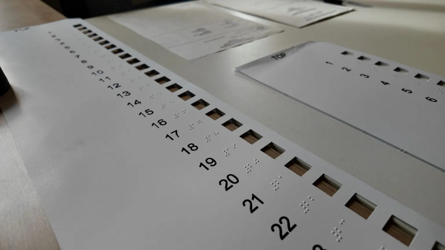A longer overlay for the bigger ballot paper has more than 20 numbers, top to bottom with Braille beside the numbers to help the voter. The overlay is white and the numbers are in black.