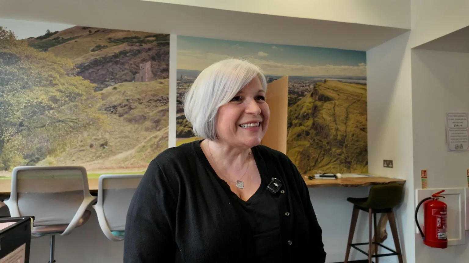 Julie Wright, a smiley woman with a white bob stands in front of the mock-up polling booth where staff are trained. She wears a black top and cardigan.