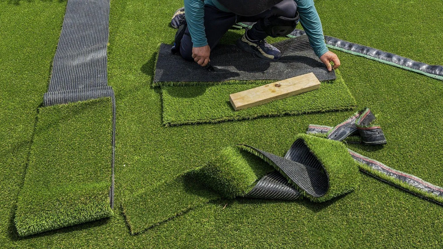 Miami Herald/ Sheets of artificial grass in a garden. There are off-cuts on top of them as well as a person, whose face cannot be seen, crouching down cutting a piece of the plastic grass. 