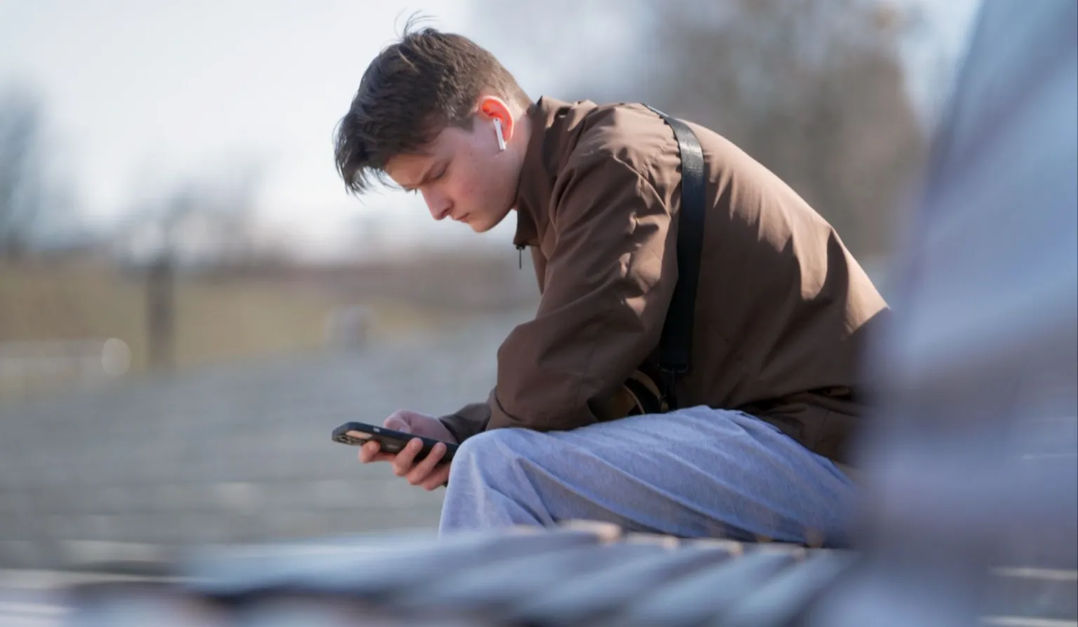A young man is seen using his mobile phone while sitting on a park bench on a clear day in Vladimir