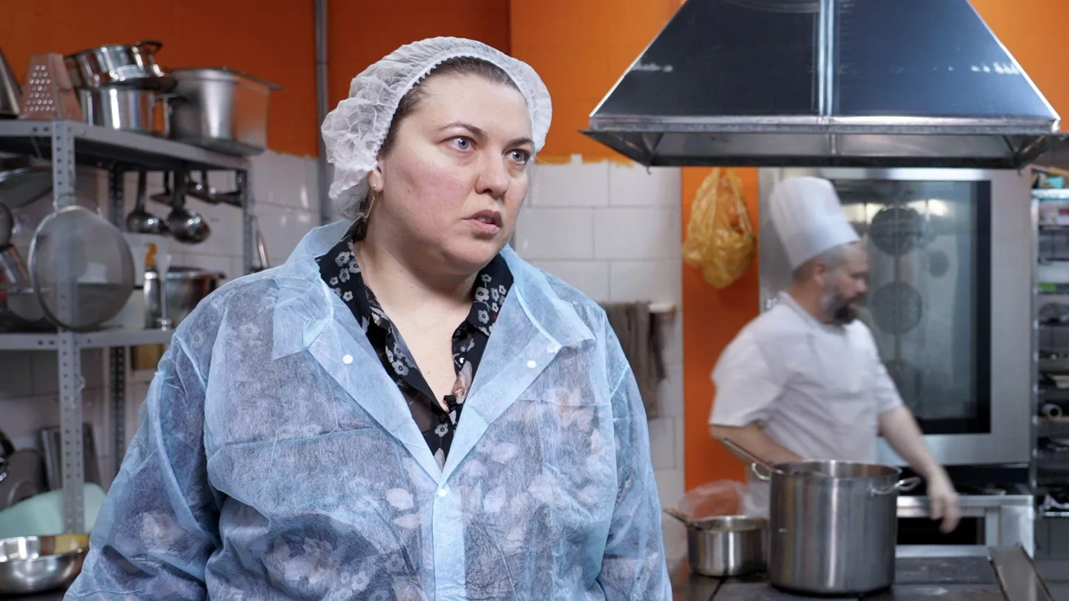 Yulia wears overalls and a hairnet in am industrial kitchen with orange walls. A man in a chefs hat and apron walks behind her. 