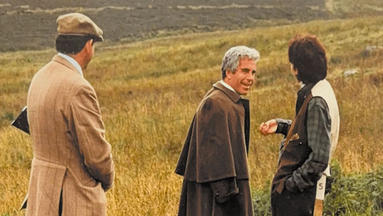 US Department of Justice Jeffrey Epstein, wearing what appears to be a cape in brown wool or tweed, turning towards a woman with short brown hair, who resembles Ghislaine Maxwell. To the left is a man in a light brown tweed jacket and a flat cap, with the stock of a shotgun tucked under his arm. Behind them is a hilly landscape with bracken and dry grass. 