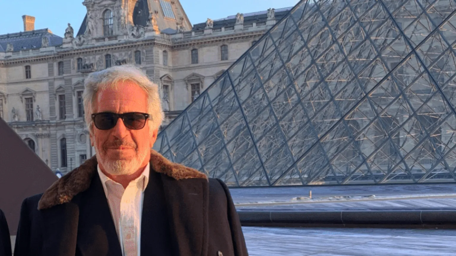 US Department of Justice Jeffrey Epstein, with a grey beard and wearing a fur-trimmed coat draped over his shoulders and sunglasses, stands in front of the Louvre and its glass pyramid.